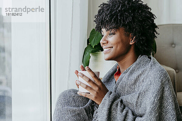 Glückliche junge Frau mit Kaffeetasse sitzt zu Hause am Fenster