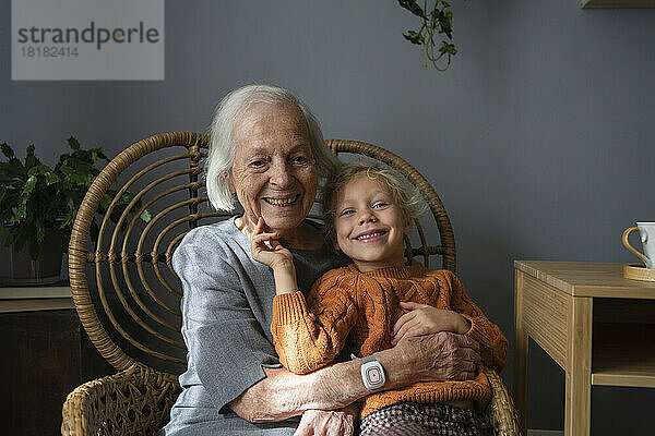 Glückliche Großmutter umarmt Enkelin zu Hause auf einem Stuhl