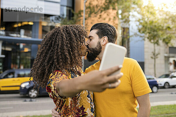 Junges Paar küsst sich und macht ein Selfie mit dem Smartphone am Fußweg