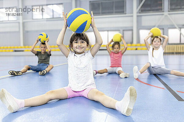 Lächelndes Mädchen hält Ball über sich und sitzt mit Freunden auf dem Schulsportplatz