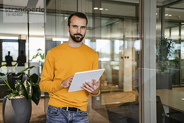 Selbstbewusster Geschäftsmann mit Tablet-PC  der im Büro an einer Glaswand lehnt