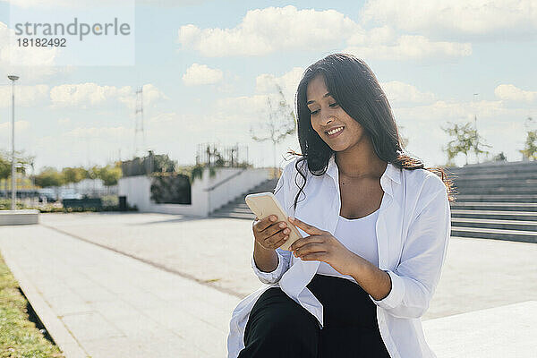 Glückliche junge Frau  die an einem sonnigen Tag im Freien sitzt und ihr Smartphone benutzt