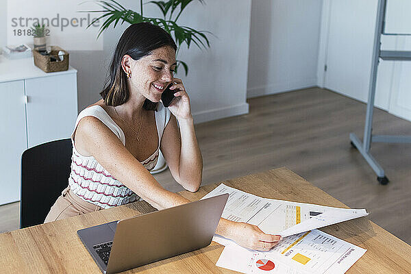 Glückliche junge Geschäftsfrau  die über ihr Smartphone spricht und sich im Büro Diagramme ansieht