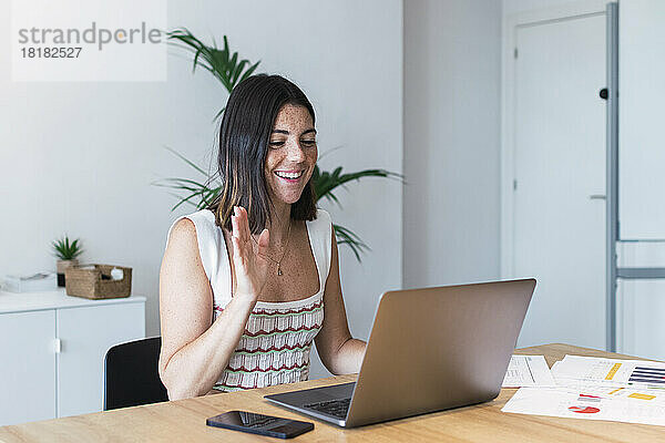 Lächelnde junge Geschäftsfrau winkt per Videoanruf dem Laptop zu
