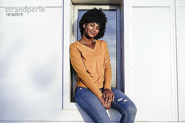 Lächelnde junge Frau mit Afro-Haaren sitzt auf der Fensterbank
