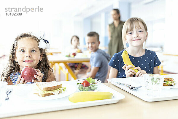 Mädchen mit Apfel und Banane sitzen am Tisch in der Cafeteria