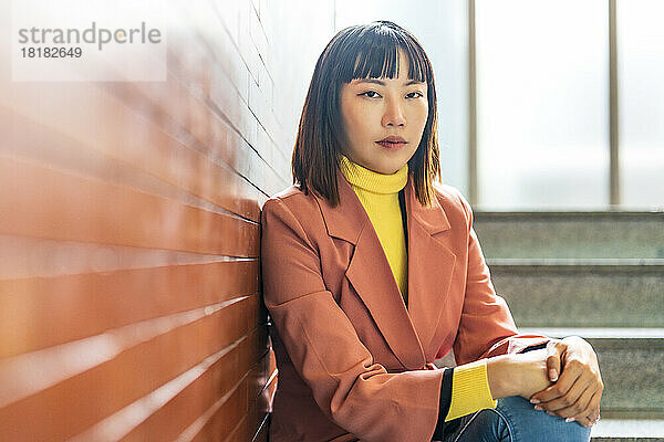 Confident young woman with bangs sitting by wall