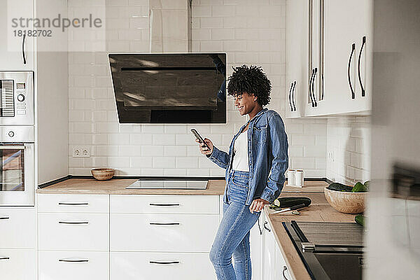 Glückliche Frau benutzt Smartphone und lehnt sich zu Hause an die Küchentheke