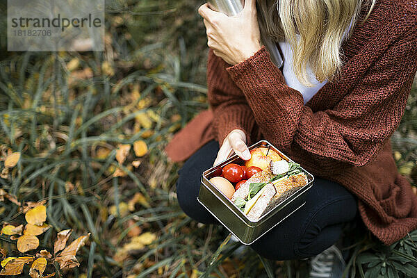 Reife Frau mit Lunchbox kauert im Herbstpark