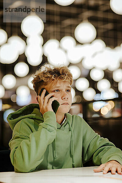 Junge spricht mit Smartphone vor Lichtern im Food-Court