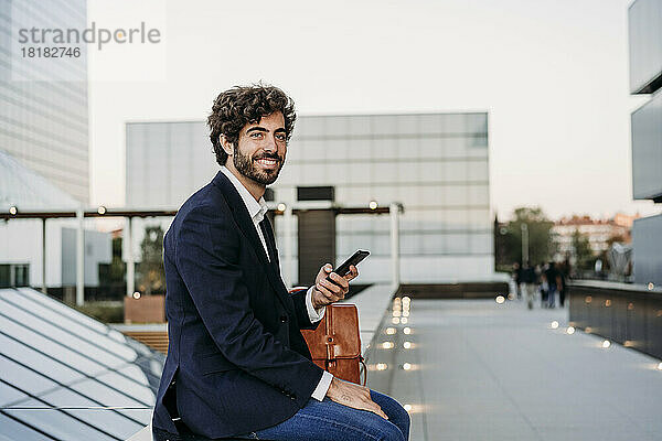 Nachdenklicher Geschäftsmann mit Smartphone an der Wand sitzend