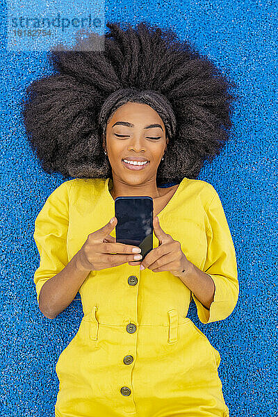 Lächelnde Frau mit Afro-Frisur benutzt Handy und liegt auf blauem Basketballplatz