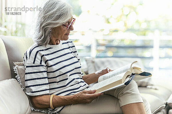 Frau liest ein Buch und sitzt zu Hause auf dem Sofa