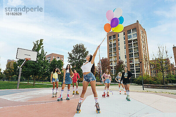 Glückliche Frau mit Rollschuhen und bunten Luftballons vor Freunden auf dem Sportplatz