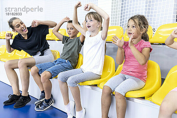 Lehrer jubeln mit Schülern  die auf dem Schulsportplatz sitzen