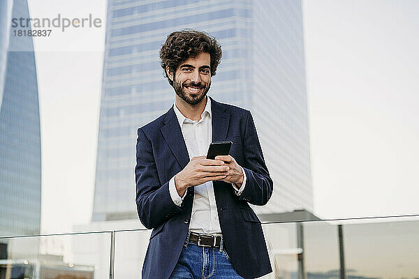 Lächelnder urbaner Geschäftsmann mit Smartphone vor dem Gebäude