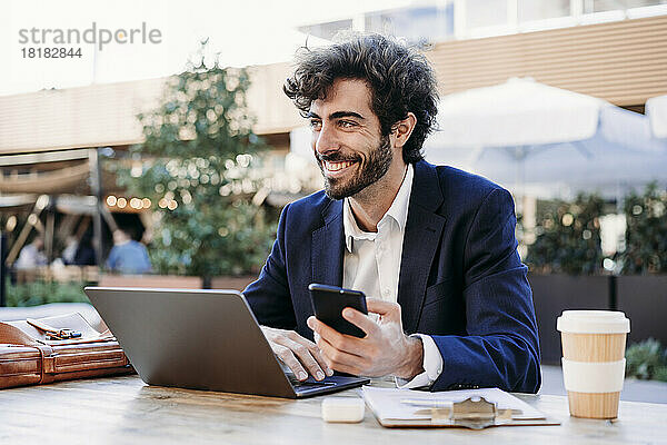 Lächelnder Geschäftsmann mit Smartphone und Laptop  der im Café arbeitet
