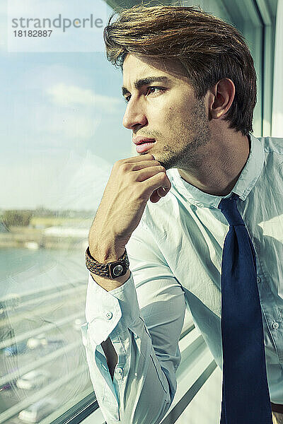 Porträt eines jungen Geschäftsmannes mit der Hand am Kinn  der durch das Fenster schaut