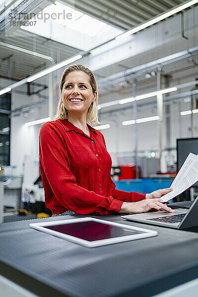 Nachdenkliche Geschäftsfrau mit Dokument und Laptop in der Fabrik