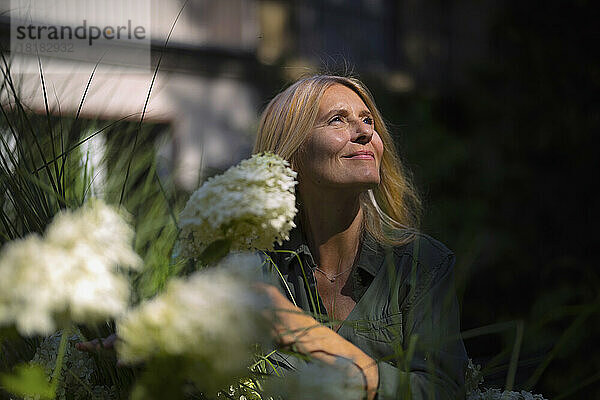 Lächelnde reife Frau steht neben blühender Pflanze im Garten