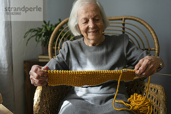 Lächelnde ältere Frau  die zu Hause einen Schal auf einem Stuhl strickt