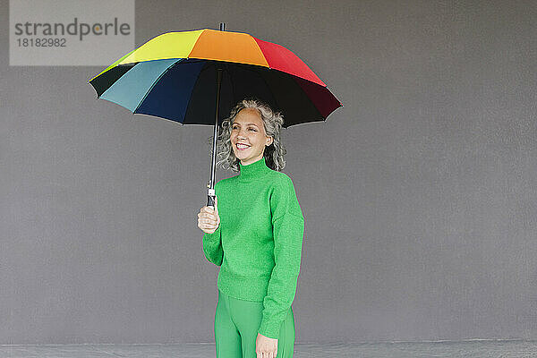Glückliche Frau mit buntem Regenschirm  die vor der Wand steht