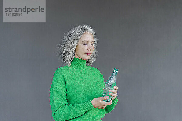 Frau mit grauen Haaren hält grüne Wasserflasche vor Wand