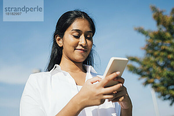 Lächelnde Frau  die Textnachrichten über das Mobiltelefon unter blauem Himmel sendet