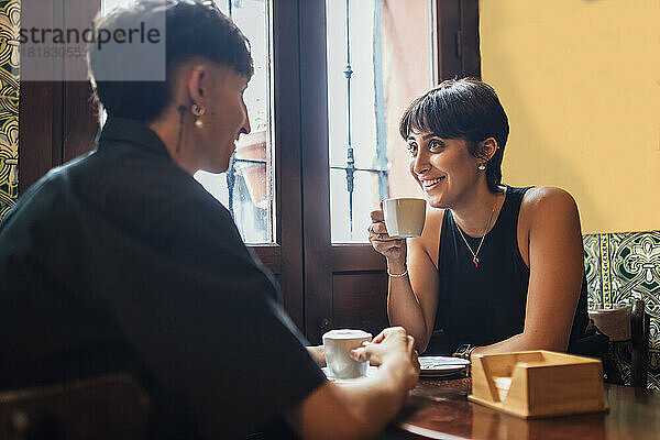 Glückliche junge Frau hält Kaffeetasse in der Hand und unterhält sich mit ihrem Freund im Café
