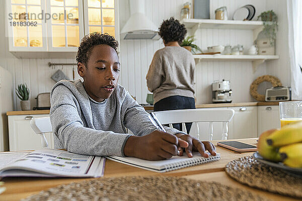 Junge schreibt an Buch  während Mutter im Hintergrund zu Hause arbeitet