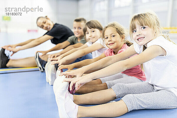 Lächelnde Grundschüler machen Übungen und sitzen Seite an Seite mit dem Lehrer auf dem Schulsportplatz