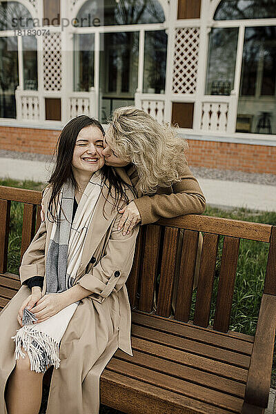 Mother kissing daughter sitting on wooden bench