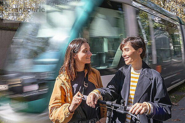 Lächelnde Frau blickt auf einen Freund  der mit einem Tretroller an der Straßenbahn vorbeifährt
