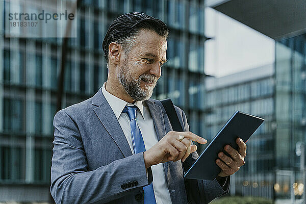 Lächelnder reifer Geschäftsmann mit Tablet-PC vor dem Gebäude