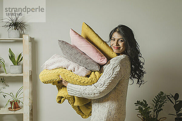 Smiling young woman holding pillows and blankets by wall at home