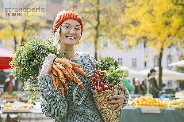 Glückliche Frau hält Gemüsebeutel in der Hand und trägt ein Bündel Karotten auf der Schulter auf dem Markt
