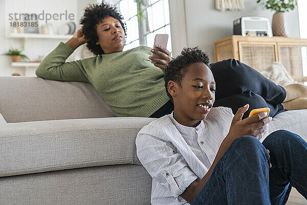 Lächelnder Junge benutzt Smartphone mit Mutter im Hintergrund zu Hause