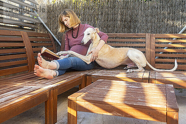 Schwangere Frau sitzt mit Hund und liest Buch auf Holzstühlen