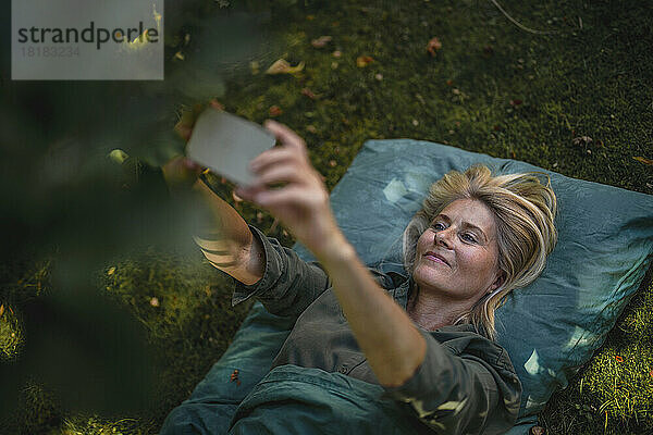 Mature woman lying on grass and taking selfie through smart phone in garden