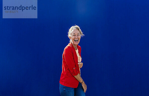 Reife Frau im roten Blazer lacht neben blauer Wand