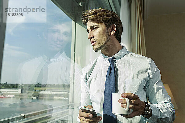 Porträt eines jungen Geschäftsmannes mit Smartphone und Tasse  der durch das Fenster schaut