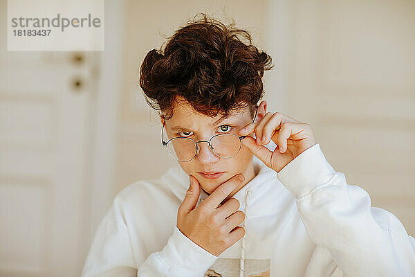 Boy adjusting eyeglasses with hand on chin at home