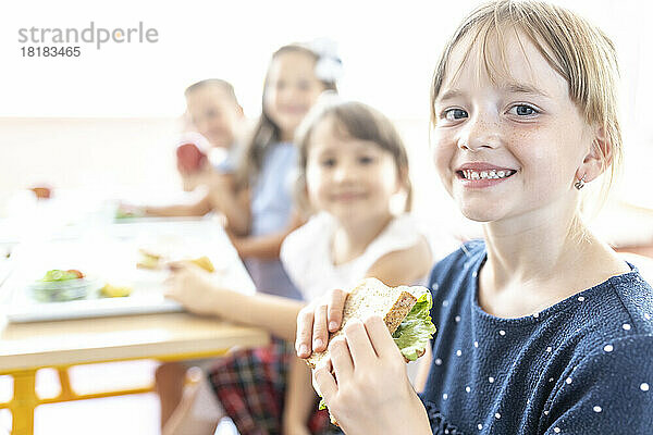 Student hält frisches Sandwich mit Freunden im Hintergrund in der Cafeteria