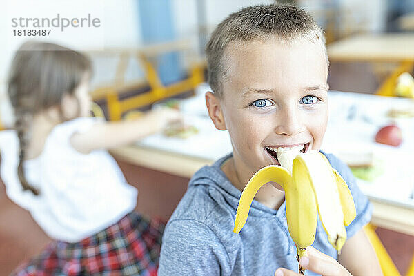 Fröhlicher Junge mit blauen Augen  der in der Mittagspause in der Cafeteria Banane isst