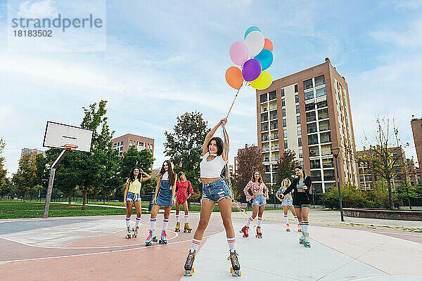 Lächelnde Frau mit Rollschuhen und erhobenen Armen hält bunte Luftballons vor Freunden auf dem Sportplatz