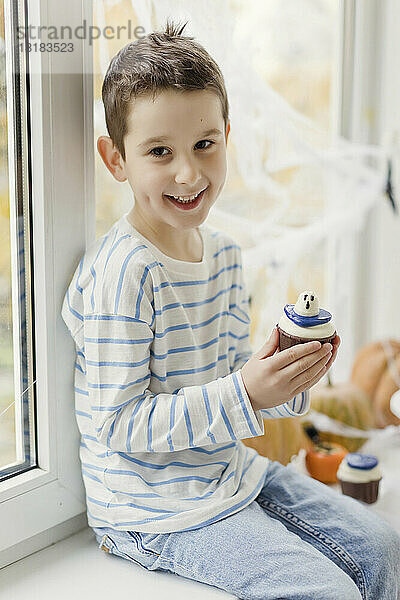 Glücklicher Junge mit Cupcake  der zu Hause auf der Fensterbank sitzt