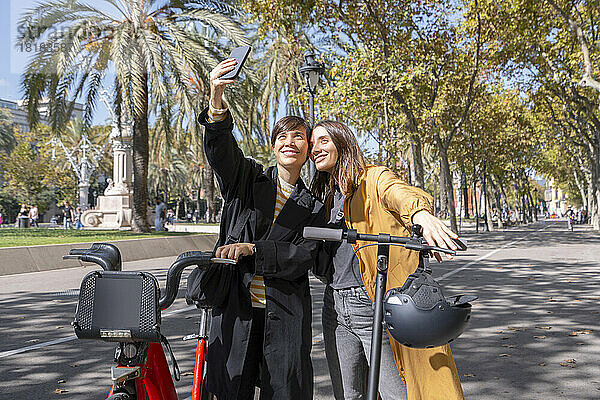 Freunde mit Elektrofahrrad und Tretroller machen Selfie mit dem Handy auf dem Fußweg