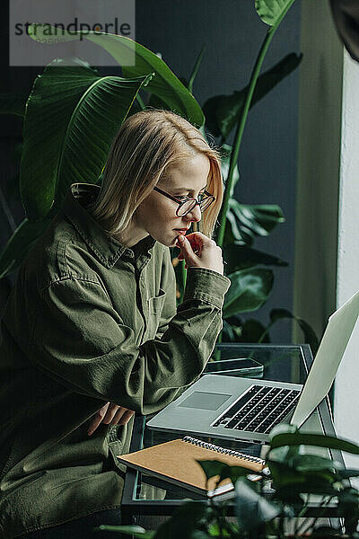 Geschäftsfrau mit der Hand am Kinn blickt im Heimbüro auf den Laptop