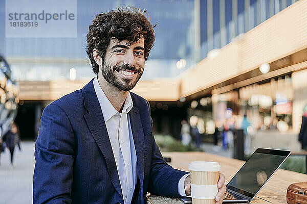 Lächelnder Geschäftsmann mit Laptop und Einwegkaffeetasse sitzt im Café