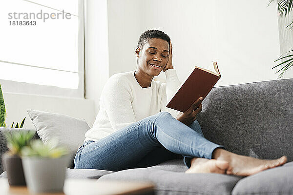 Lächelnde Frau mit geschlossenen Augen sitzt zu Hause auf der Couch und hält ein Buch in der Hand